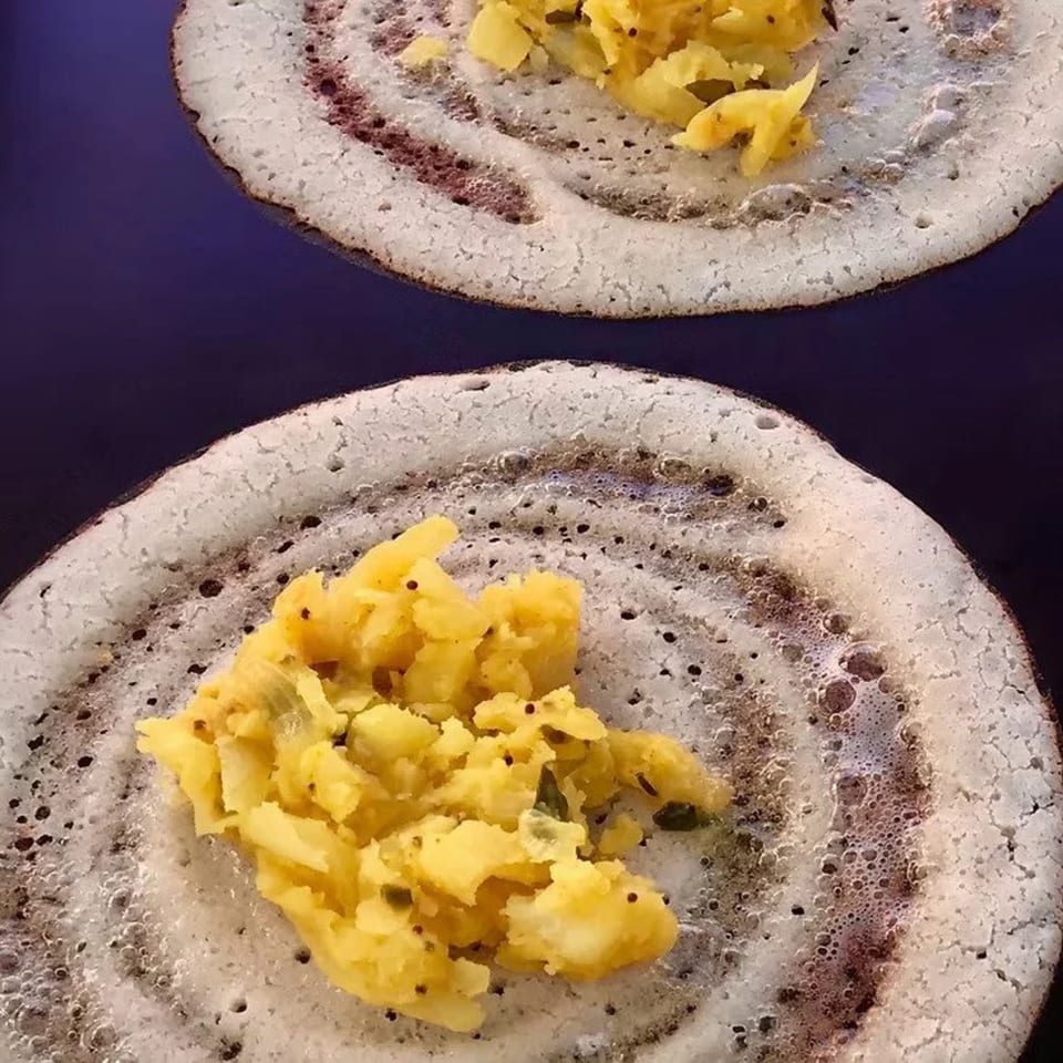 Two dosas, light brown and bubbly, cooking on a dark surface with potato filling.