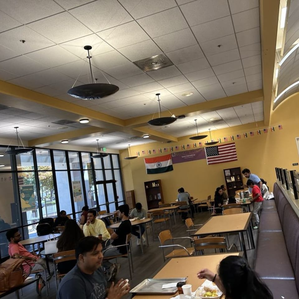 A cafeteria with students eating at tables, an Indian flag, and an American flag on the wall.
