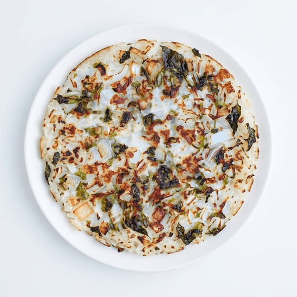 A round, savory pancake with charred onions and green herbs on a white plate.