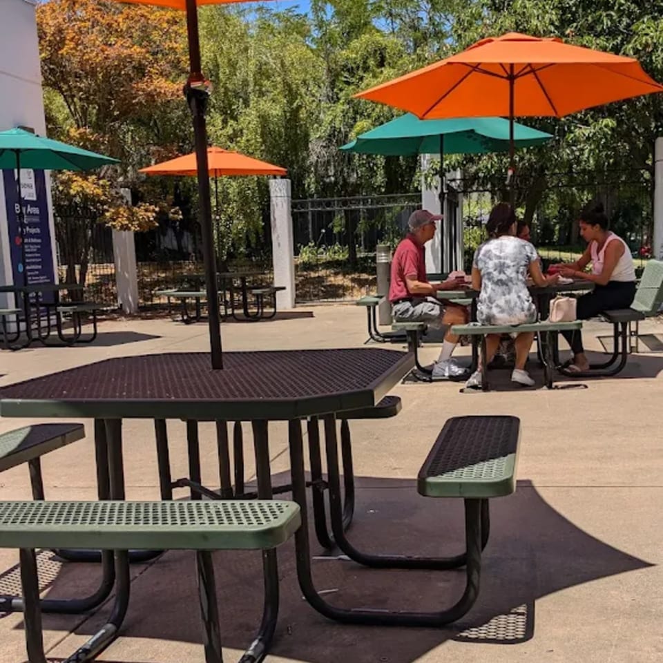 People dining outdoors at picnic tables with orange and green umbrellas providing shade.