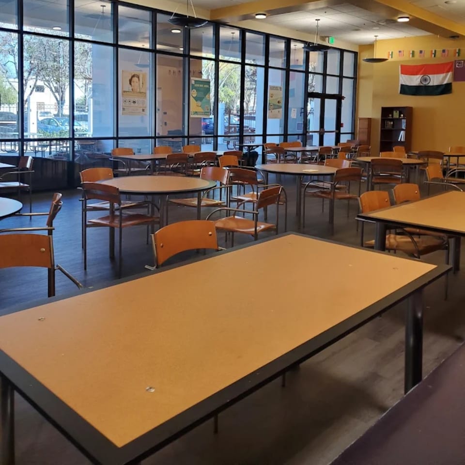 Empty dining hall with many tables and chairs, large windows, and an Indian flag on the wall.